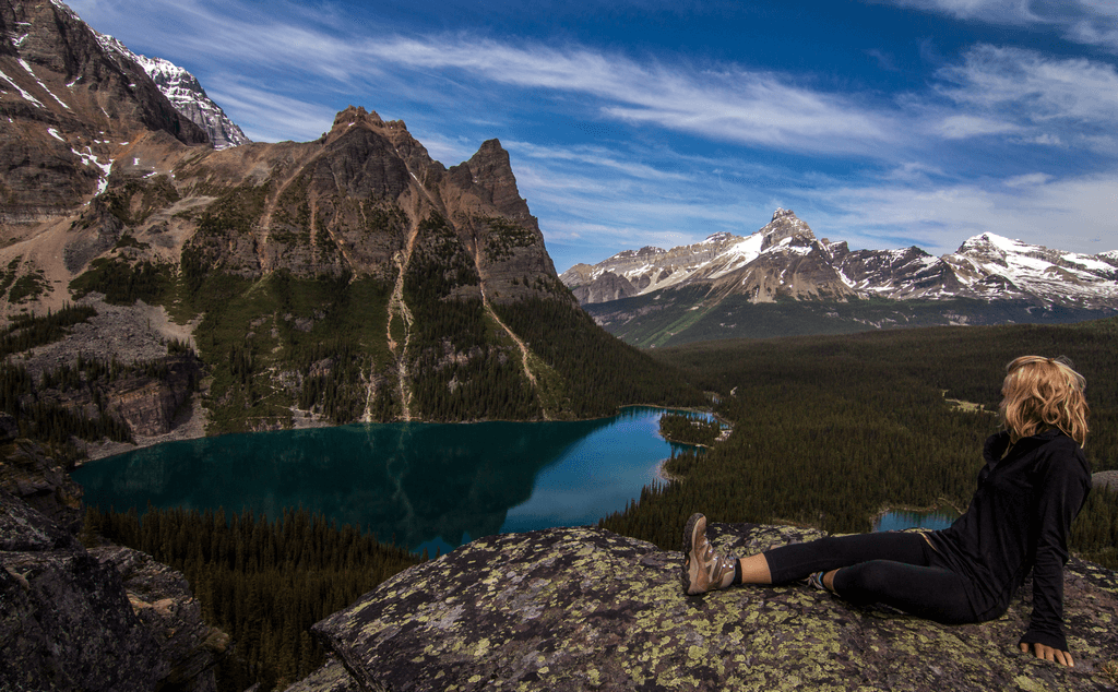 Wandern in Kanada. Eine junge Frau sitzt mit Wanderschuhen auf einem Felsen. Im Hintergrund sind Berge zu sehen, die teilweise mit Schnee bedeckt sind. Auch ist ein See zu sehen.